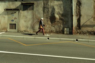 An old woman walking down a street with old buildings but a fresh road