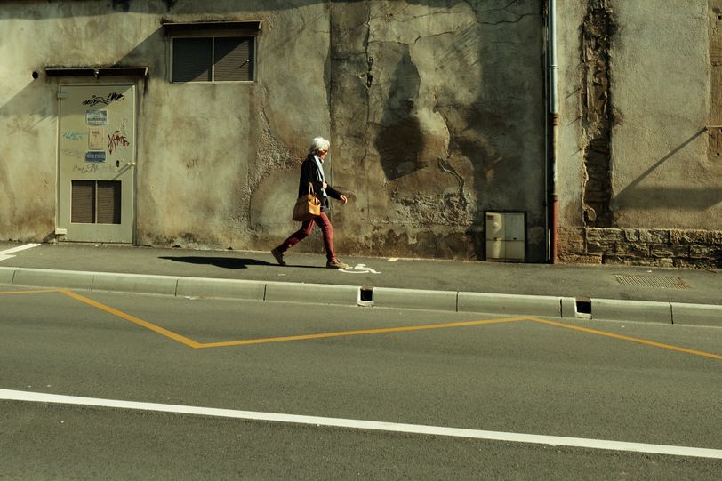 An old woman walking down a street with old buildings but a fresh road