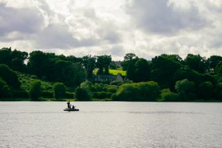 View on a lake with the surface of the lake and people on a boat, houses and trees in the back and a blue sky