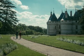 Two people walking a dog on a path coming from a gigantic renaissance castle