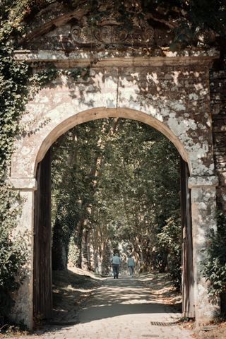 Massive door made of stone opening to a straight path under multiple trees where some people are walking