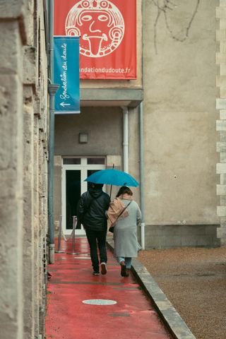 A couple with a blue umbrella walking on a red footpath to a building decorated with blue and red signs