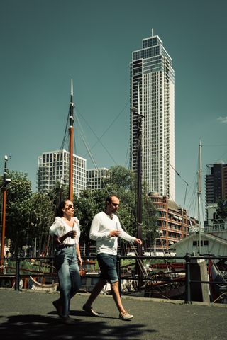 Two people walking side by side on a marina with boats behind them and two big skycrapers side by side in the background