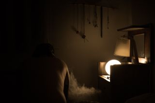 A man sitting on a bed in the a dark bedroom with only one source of light