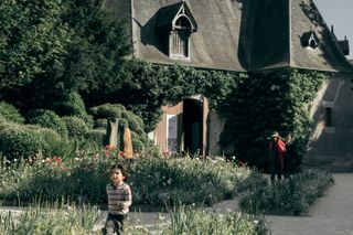 View of a house in a park with a little boy running in the foreground and his guardians walking behind him while keeping an eye on him.