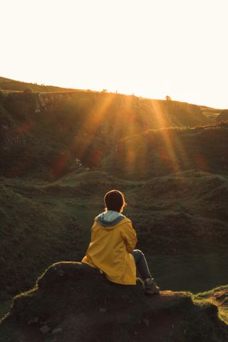 The back of a person sit on a rock in the foreground facing the setting sun and a moutain in the background