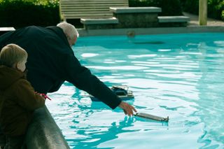 A grand-father is helping his grand-son by trying to refloat his miniature boat in a pond