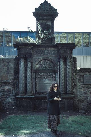 Young woman with a phone in her hands stepping away from an ancient massive headstone in a cemetery