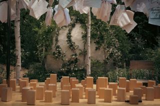 Close view to a wooden table with wooden blocks displayed evenly on it. Above the table are hanging several white sheets of paper. And a white wall covered in plants in the background.