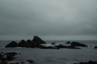 Several rocks emerging from the sea where dozens of birds land to rest on a very cloudy day.