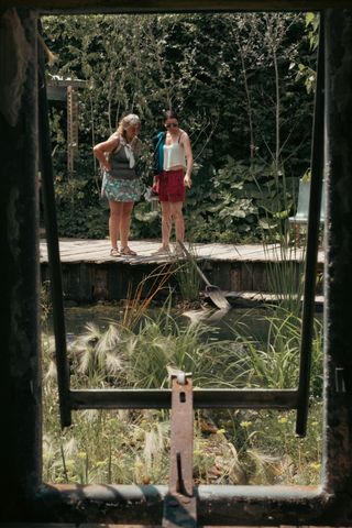 Seeing through a frame two woman are looking down on a pond with one of them pointing something in it