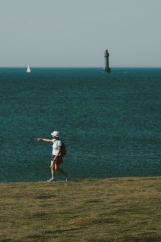 A woman on a cliff pointing out of the frame with a sailing boat and a lighthouse in the background