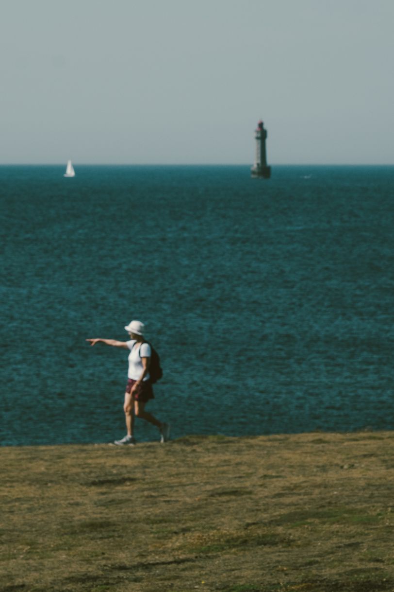 A woman on a cliff pointing out of the frame with a sailing boat and a lighthouse in the background