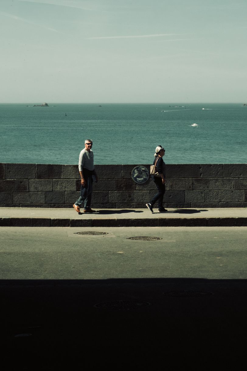 A couple walking on a road next to the sea with layers of light, colors and shadow