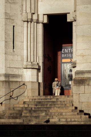 An old man sitting before a church's door and a pigeon down the stairs watching each other