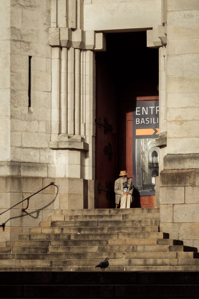 An old man sitting before a church's door and a pigeon down the stairs watching each other