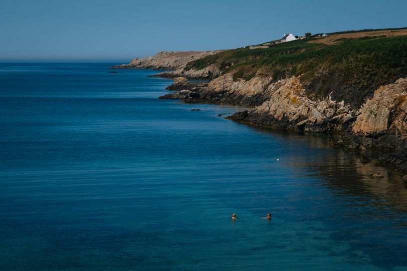 Two people swimming in a blue calm sea next to a cliff