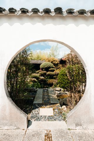 A round gate in a white wall to a flourished asian garden
