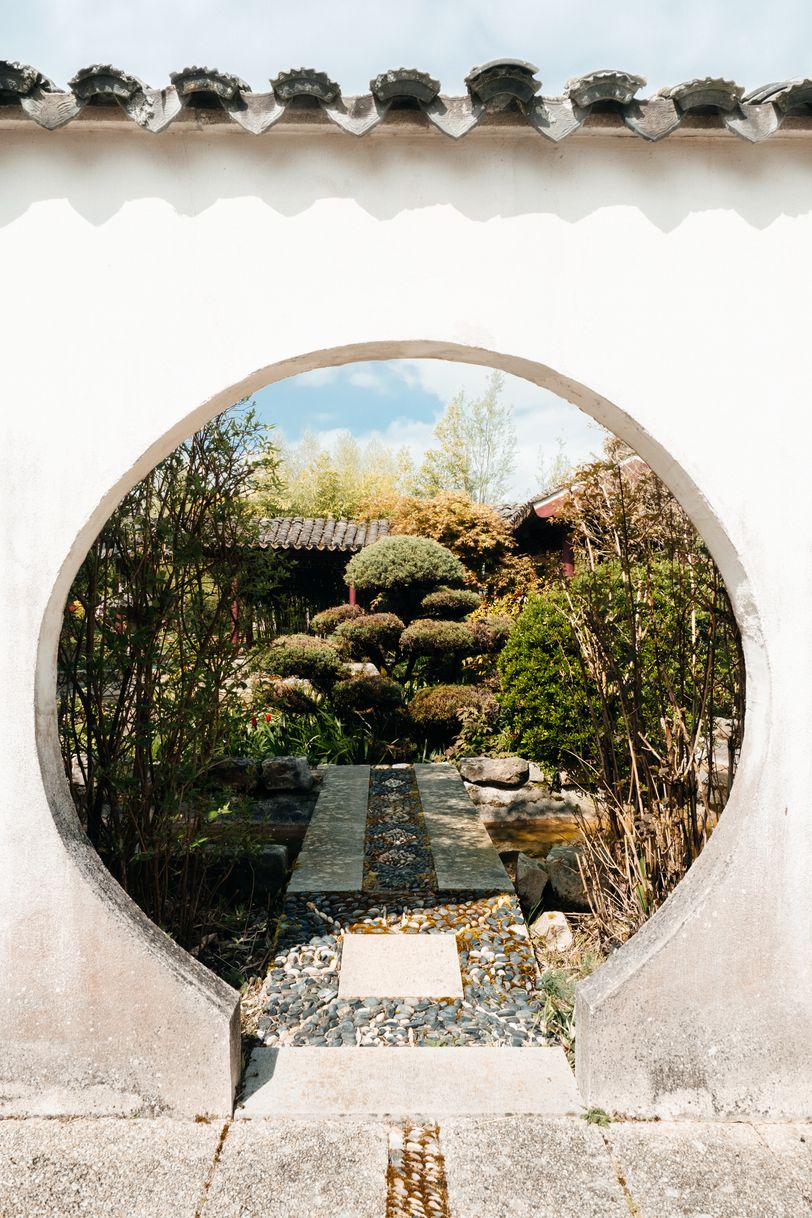 A round gate in a white wall to a flourished asian garden